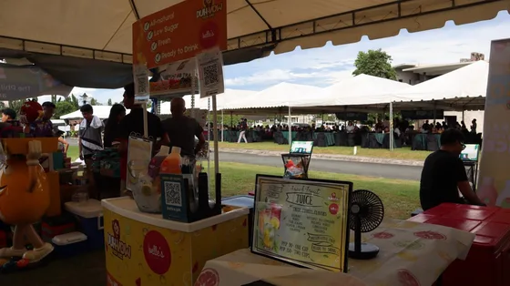 A juice stall counter at the pop-up with the stall's printed bXCODE QR card displayed above it and a bXTRA sign-up QR stand on the counter, with the market and seated crowd behind.
