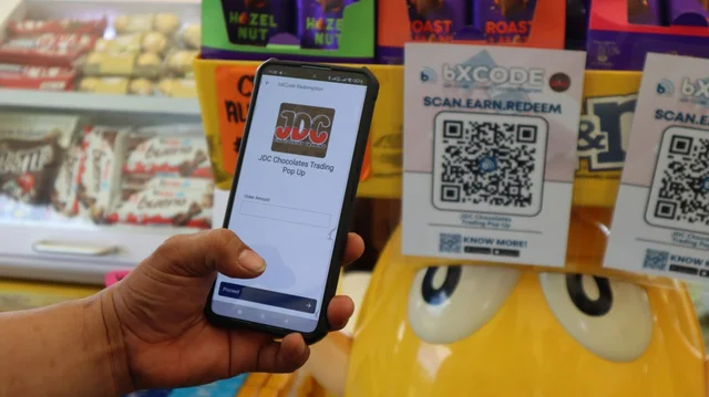 A bXTRA tent at the pop-up with a large bXCODE QR displayed on the counter panel and a staff member seated behind it.