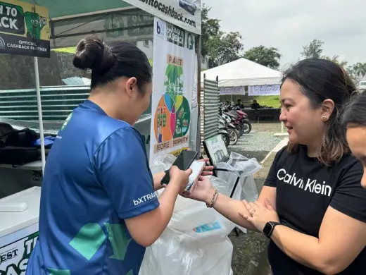 A booth attendant scanning a customer's phone at the exchange counter, with bagged recyclables on the ground beside them.