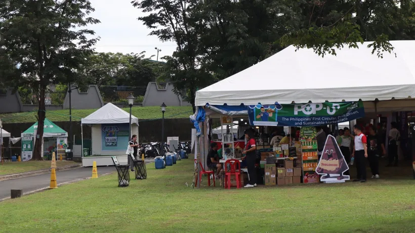 The pop-up seen from across the lawn: a row of white marquees over food stalls with a banner reading
         "Pop-Up — The Philippines' First Circular, Data-Driven, and Sustainable Marketplace", the green
         Trash to Cashback exchange tents at the far end, and visitors walking between them.