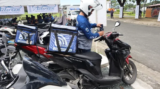 Two bXTRA Community Rider insulated delivery boxes mounted on motorbikes at the pop-up, with a rider in a bXTRA jersey and helmet beside them and the event's banners behind.