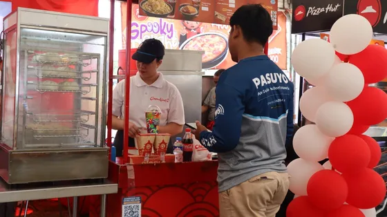 A PASUYO rider in a bXTRA-branded jersey collecting a bagged order at a food stall counter during the pop-up, with a bXTRA app QR card on the counter and other stalls behind.