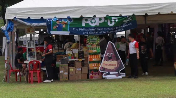 A stall trading under a marquee at the pop-up: the stallholder at her counter with shelves of drinks and snacks, a banner above reading "Pop-Up — The Philippines' First Circular, Data-Driven, and Sustainable Marketplace", and visitors walking past on the grass.