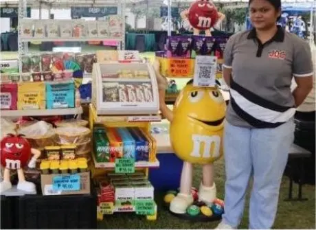 A stallholder standing at her counter, shelves of snacks and drinks priced and laid out in front of her.